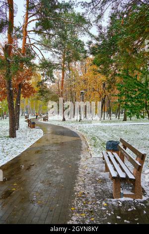 Erster Schneefall im bunten Stadtpark im Herbst. Einsame Bank auf Gasse unter Bäumen brabches mit goldenen, grünen, orange Laub weißen Schnee covere Stockfoto