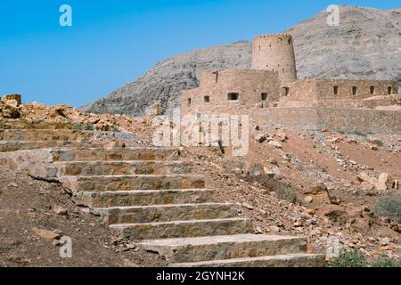 Steinmauern einer kleinen mittelalterlichen arabischen Festung unter hohen Bergklippen. Festung in Bukha, Halbinsel Musandam, Oman. Heißer, dunstiger Tag in der arabischen Wüste. Stockfoto