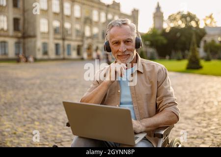 Porträt eines glücklichen, reifen Mannes, der im Rollstuhl einen Patienten mit Kopfhörern und lächelnder Kamera erholt und im Park in der Nähe der Klinik einen Laptop benutzt Stockfoto