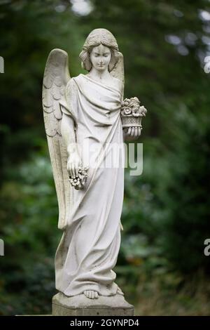 Weißer Engel mit Blumen in der Hand vor verschwommenem grünen Hintergrund auf einem Friedhof Stockfoto