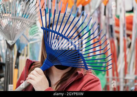 Frau im Geschäft kauft Besen, um abgefallene Blätter zu sammeln. Sie trägt eine Maske Stockfoto