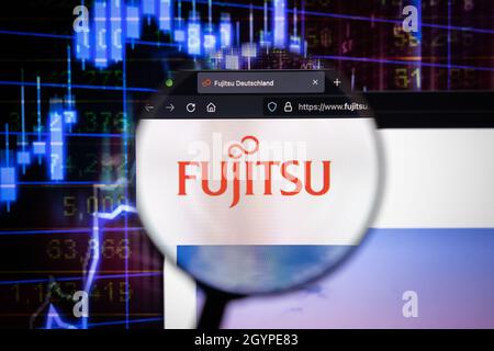 Fujitsu-Firmenlogo auf einer Website mit verschwommenen Börsengrafiken im Hintergrund, die auf einem Computerbildschirm durch eine Lupe gesehen werden. Stockfoto