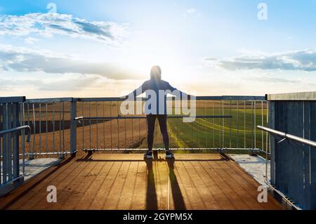 Der junge Sportler beobachtet die landwirtschaftlichen Felder über den Brückengeländer. Dramatische Abendszene, Strahlen der untergehenden Sonne. Stockfoto