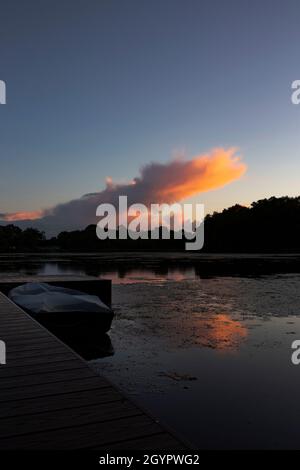 Das Boot vertäute bei Sonnenuntergang an einem Steg auf einem See. Mit Wald im Hintergrund, der sich im Wasser widerspiegelt Stockfoto