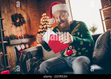 Foto von aufgeregt froh beeindruckt Kerl offen umhüllen nehmen Brief tragen weihnachtsmann Hut Pullover dekoriert Haus drinnen Stockfoto