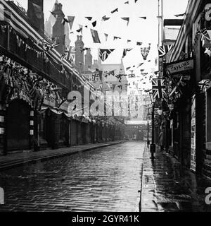 Schilder für „Fish & Chips“ und einen Kutschstand auf einer typischen Straße in einer englischen Marktstadt in den 1930er Jahren - The Shambles, Huddersfield, Yorkshire, dekoriert zur Feier der Krönung des britischen Monarchen König George VI. Am 12. Mai 1937. Vintage-Foto aus einer privaten Sammlung. Stockfoto