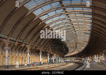 Ein Panorama einer Bahnhofskonkurse. Ein eiserner Baldachin aus dem 19. Jahrhundert zieht sich über die Plattformen und das Sonnenlicht fällt auf die Plattform. Eine Fußbrigge Stockfoto