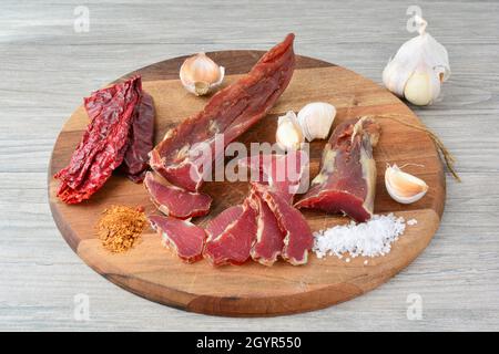 Geräuchertes Schweinefleisch auf dunklem Holzbrett, mit ein paar trockenen roten Paprika, etwas Knoblauch, Salz und würziger scharfer Chilischote arrangiert Stockfoto