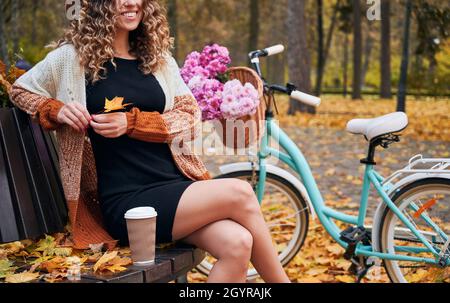 Elegante junge Frau mit Herbstblatt und Lächeln auf der Bank mit einer Tasse Kaffee in der Nähe des Fahrrads. Fröhliche weibliche Person, die sich im Freien in der Nähe des Fahrrads mit Blumen im Korb ausruhte. Stockfoto