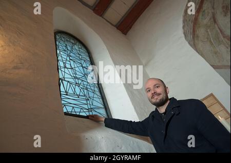 Sachsen Anhalt, Deutschland. Oktober 2021. 09. Oktober 2021, Sachsen-Anhalt, Altjeßnitz: Der Künstler Bastian Muhr steht neben einem der modernen Kirchenfenster, die er in der kleinen romanischen Kirche im Landschaftspark Altjeßnitz entworfen hat. Diese neuen Fenster sind Teil des Projekts „Lichtungen“ der Evangelischen Landeskirche Anhalt, das zeitgenössische Glasmalerei in die historischen Kirchen Anhalts bringt. Die Fenster wurden bereits im vergangenen Jahr mit Unterstützung der Landeskirche Anhalt und der Ecclesia Foundation installiert, konnten aber aufgrund der Corona-Pandemie bis jetzt nicht eingeweiht werden. Foto: Heiko Stockfoto