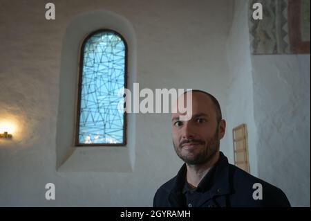 Sachsen Anhalt, Deutschland. Oktober 2021. 09. Oktober 2021, Sachsen-Anhalt, Altjeßnitz: Der Künstler Bastian Muhr steht neben einem der modernen Kirchenfenster, die er in der kleinen romanischen Kirche im Landschaftspark Altjeßnitz entworfen hat. Diese neuen Fenster sind Teil des Projekts „Lichtungen“ der Evangelischen Landeskirche Anhalt, das zeitgenössische Glasmalerei in die historischen Kirchen Anhalts bringt. Die Fenster wurden bereits im vergangenen Jahr mit Unterstützung der Landeskirche Anhalt und der Ecclesia Foundation installiert, konnten aber aufgrund der Corona-Pandemie bis jetzt nicht eingeweiht werden. Foto: Heiko Stockfoto