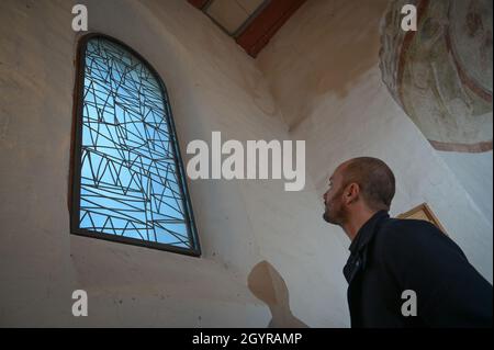 Sachsen Anhalt, Deutschland. Oktober 2021. 09. Oktober 2021, Sachsen-Anhalt, Altjeßnitz: Der Künstler Bastian Muhr blickt auf eines der modernen Kirchenfenster, die er in der kleinen romanischen Kirche im Landschaftspark Altjeßnitz entworfen hat. Diese neuen Fenster sind Teil des Projekts „Lichtungen“ der Evangelischen Landeskirche Anhalt, das zeitgenössische Glasmalerei in historische Kirchen Anhalts bringt. Die Fenster wurden bereits im vergangenen Jahr mit Unterstützung der Landeskirche Anhalt und der Ecclesia Foundation installiert, konnten aber aufgrund der Corona-Pandemie bis jetzt nicht eingeweiht werden. Foto: Heiko Stockfoto