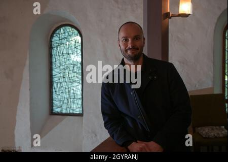 Sachsen Anhalt, Deutschland. Oktober 2021. 09. Oktober 2021, Sachsen-Anhalt, Altjeßnitz: Der Künstler Bastian Muhr steht neben einem der modernen Kirchenfenster, die er in der kleinen romanischen Kirche im Landschaftspark Altjeßnitz entworfen hat. Diese neuen Fenster sind Teil des Projekts „Lichtungen“ der Evangelischen Landeskirche Anhalt, das zeitgenössische Glasmalerei in die historischen Kirchen Anhalts bringt. Die Fenster wurden bereits im vergangenen Jahr mit Unterstützung der Landeskirche Anhalt und der Ecclesia Foundation installiert, konnten aber aufgrund der Corona-Pandemie bis jetzt nicht eingeweiht werden. Foto: Heiko Stockfoto