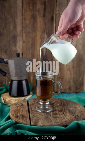 Trinken Sie in einer Tasse. Die Milch wird in den Dekanterkaffee gegossen. Geyser Kaffeemaschine, Milchpulver auf einem Holztisch. Tasse aus Glas mit Cappuccino. Kaffee zubereiten, Hand aus nächster Nähe. Stockfoto