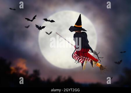 Kleine Hexe fliegt Besen an Halloween-Nacht. Riesiger Vollmond und Fledermäuse im Hintergrund. Kinder Trick or Treat Kostüm. Viel Spaß. für Kinder Gruselig Stockfoto