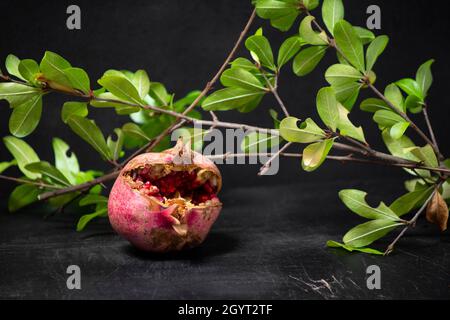 Weicher Fokus aus rotem Granatapfel und Samen mit grünen Blättern auf altem Holzschneidebrett, Seitenansicht, dunkler Hintergrund Stockfoto