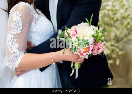 Braut und Bräutigam umarmen sich, indem sie den Brautstrauß aus weißen, gelb-rosa-violetten Blumen in die Hand nehmen Stockfoto