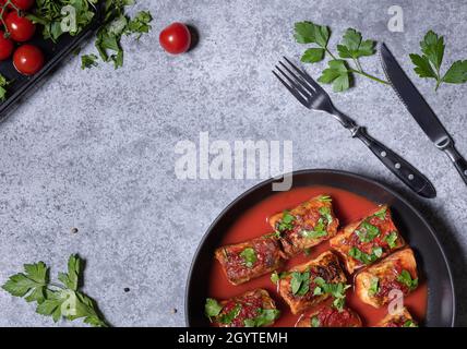 Kohlröllchen in Tomatensauce und gehackter Petersilie in der Pfanne. Flach mit Kohlrollen, einigen Zutaten, Gabel und Messer auf grauem Stein-Bac legen Stockfoto