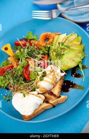 Pochiertes Ei auf Toast mit Avocado. Gemüsesalat mit lachsrotem Ryboy. Blaue Gerichte und ein Teller. Gesundes Frühstück aus nächster Nähe. Gekochte Eier und Micro Green. Stockfoto