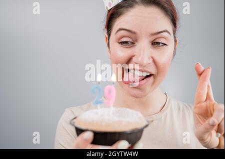 Glückliche kaukasische Frau, die ihre Zunge herausstreckte und mit gekreuzten Fingern die Kerzen auf dem Kuchen ausbläst. Das Mädchen feiert ihren 29. Geburtstag Stockfoto