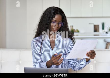 Junge schwarze Frau, die Papier in der Hand hält und im Heimbüro am Laptop arbeitet. Stockfoto