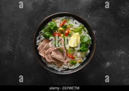 Pho Bo Suppe mit Rindfleisch, Reisnudeln, Limette, Chilischote in schwarzer Schüssel isoliert auf schwarzem Hintergrund. Blick von oben. Vietnamesische und asiatische Küche. Stockfoto