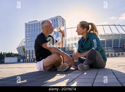 Aktives reifes Paar in Sportkleidung, reden und trinken Wasser, entspannen nach dem Training an einem sonnigen Sommertag Stockfoto
