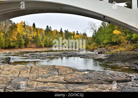 Ein Fluss mit einem niedrigen Wasserstand, der im Sommer wegen des geringen Regens über Felsen fließt und größere Felsen freilegt, während die Bäume Herbstfarben zeigen. Stockfoto