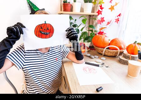 Ein Kind zeigt seine Zeichnung zu Halloween, an einem Schreibtisch am Fenster sitzend, umgeben von Kürbissen und Zimmerpflanzen. Skizze eines orangen Lachens Stockfoto