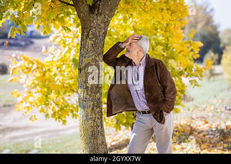 Ein reifer Mann mit grauem Haar steht an einem Baumstamm gelehnt. Er hat Ärger oder Müdigkeit. Stockfoto