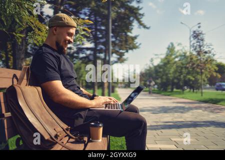 Ein positiver Mann mit Laptop, der draußen auf der Bank im Stadtpark arbeitet. Distanzjob Stockfoto