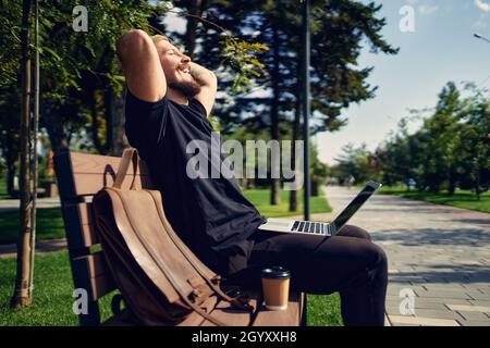 Der junge Millennial-Hipster sitzt mit einem Laptop auf der Bank im Park und genießt die Entspannung in der Freizeit Stockfoto