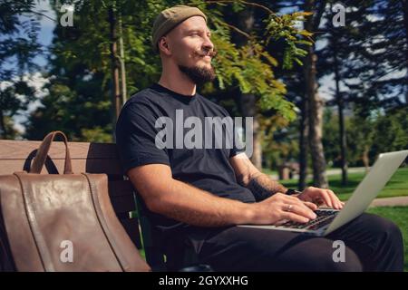 Der junge kaukasische Freiberufler sitzt mit einem Laptop auf der Bank im Park und genießt eine entspannte Freizeit Stockfoto