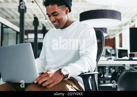 Lächelnder Geschäftsmann, der in einem modernen Büro an einem Laptop arbeitet. Glücklicher junger Geschäftsmann, der gerne allein an einem modernen Arbeitsplatz arbeitet. Kreative junge Busin Stockfoto