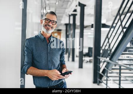Glücklicher, reifer Geschäftsmann, der ein Smartphone hält und die Kamera anlächelt. Kreativer Geschäftsmann, der allein an einem modernen Arbeitsplatz steht. Erfahrene Busine Stockfoto