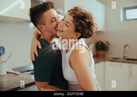 Queeres Paar lacht fröhlich in der Küche. Zwei romantische junge queere Liebhaber umarmen sich gegenseitig, während sie zusammen stehen. Fröhlicher junger LGBTQ+ c Stockfoto