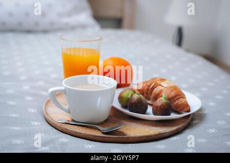 Frühstück im Bett. Croissant auf einem Teller mit Feigen, einer Kaffeetasse, Orangensaft und einer Orange auf einem Tablett Stockfoto