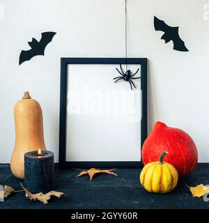 Halloween Feiertagskonzept mit leerem Bilderrahmen, Kürbissen, Kerze, Fledermäusen und Spinne auf schwarzem Tisch gegen weiße Wand. Platz kopieren, Modell erstellen. Stockfoto