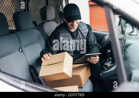 Lächelnder männlicher Kurier in Uniform und Mütze, der Informationen auf Karton überprüft, während er mit einem digitalen Tablet im Auto sitzt Stockfoto