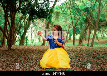 Ein süßes kleines hispanische Mädchen, das ein schneeweißes Kleid trägt und einen Apfel in einem Wald hält Stockfoto