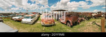 Panoramablick auf einen Oldtimer-Schrottplatz unter einem teilweise bewölkten Himmel mit verschiedenen Oldtimern und rostigen Fahrzeugen auf Gras Stockfoto