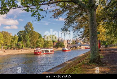 Zwei Touristenboote segeln den Fluss hinauf mit einer historischen Brücke im Hintergrund. Ein Pfad verläuft entlang des Flusses und am Flussufer befinden sich Herbstbäume. Stockfoto