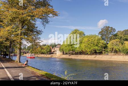 Eine Szene am Fluss im Frühherbst. Ein Touristenboot fährt flussabwärts in der Nähe eines historischen Verteidigungsturms, und eine junge Frau sitzt am Flussufer. Stockfoto