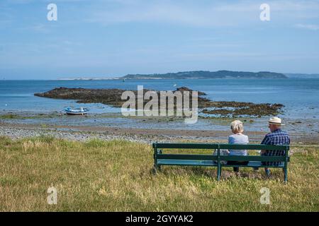 Ehepaar sitzt auf einer Bank in der Stadt mit Blick auf Herm Island, Guernsey, Channel Islands Stockfoto