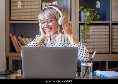 Geschäftsfrau, die im Heimbüro Musik über Kopfhörer hört, während sie am Laptop arbeitet. Kaukasische junge Frau, die Musik über Kopfhörer genießt. Fröhliche Frau Stockfoto