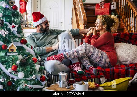 Kaukasisches Paar, das während der Weihnachtsfeier zu Hause auf dem Sofa im Wohnzimmer sitzt, Spaß hat. Paar in Hut und Socken mit geschmückter Christma Stockfoto