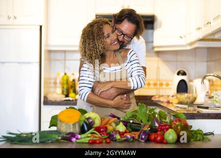 Liebevoller Mann umarmt seine Frau von hinten in der Hausküche. Mann umarmte Frau in der Küche mit einer Vielzahl von frischem rohem Gemüse auf der Küchenarbeitsfläche Stockfoto
