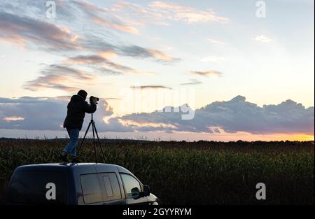 Ein Mädchen fotografiert den Sonnenuntergang von einem Stativ aus, das auf dem Dach eines Autos auf dem Feld steht. Stockfoto