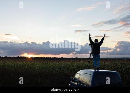 Ein Mädchen fotografiert den Sonnenuntergang von einem Stativ aus, das auf dem Dach eines Autos auf dem Feld steht. Stockfoto