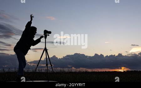 Ein Mädchen fotografiert den Sonnenuntergang von einem Stativ aus, das auf dem Dach eines Autos auf dem Feld steht und den Raum kopiert. Stockfoto